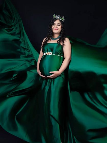 A powerful and regal portrait featuring a deep emerald green satin gown. The fabric is expertly tossed to create a wing-like effect, adding a touch of fantasy to this maternity studio photograph.