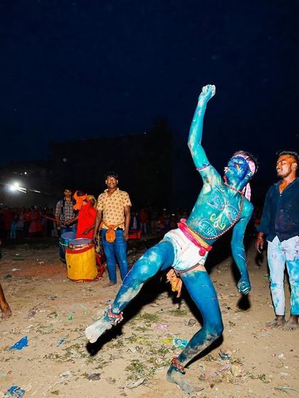 A performer painted in brilliant blue strikes a dynamic pose against the night sky during the Gajan festival, showcasing the dance and movement central to the ritual.