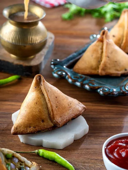The quintessential Indian snack, a samosa, presented with chai. The warm, rustic styling evokes a sense of comfort and tradition, making it a relatable and tempting image.
