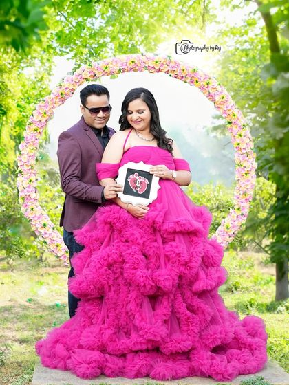 A sweet couple's portrait holding a sonogram picture, framed by a circular floral prop. The mother-to-be is wearing a vibrant pink ruffled gown.