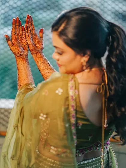 A candid shot of the bride from the back, with her hands raised in a prayer-like gesture, showing off the mehndi that extends up her arms.