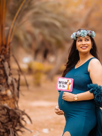 A beautiful solo portrait of a mom-to-be holding a "Mom to Be" prop. She wears a floral crown and a lovely blue gown in a natural outdoor setting.