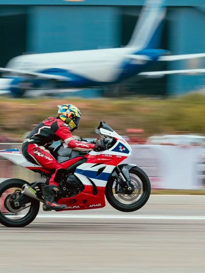 A superbike takes flight, pulling a wheelie right off the starting line with an airplane in the background. This is speed versus sky at the Taneja Aerodrome.
