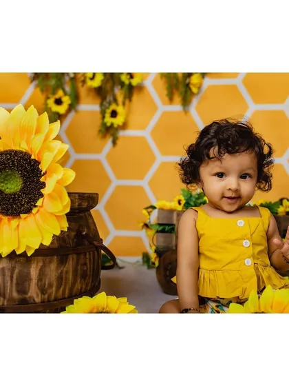 A cheerful baby girl in a yellow outfit waves at the camera, surrounded by a vibrant sunflower and honeycomb-themed studio backdrop. This setup is perfect for a bright and happy baby portrait.