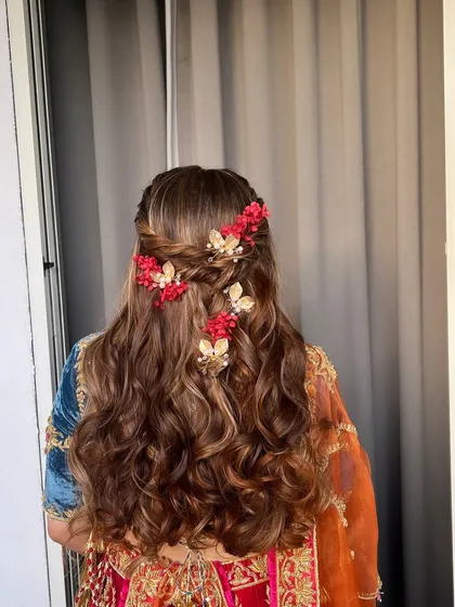 A whimsical Mehendi hairstyle with soft waves and a braided crown, adorned with red flowers and golden butterfly clips.