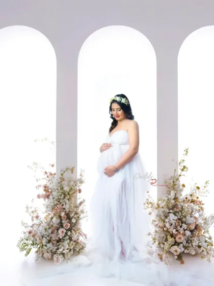 A serene and classic portrait of a mother-to-be in a sheer white gown. She stands between two floral arrangements against a clean, arched backdrop, creating an angelic look.