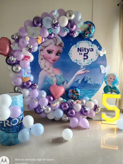 This is a popular Disney Frozen theme I created for a 5th birthday party. It includes a custom backdrop with Elsa, a balloon garland in icy blue, purple, and silver, and a light-up number '5'.