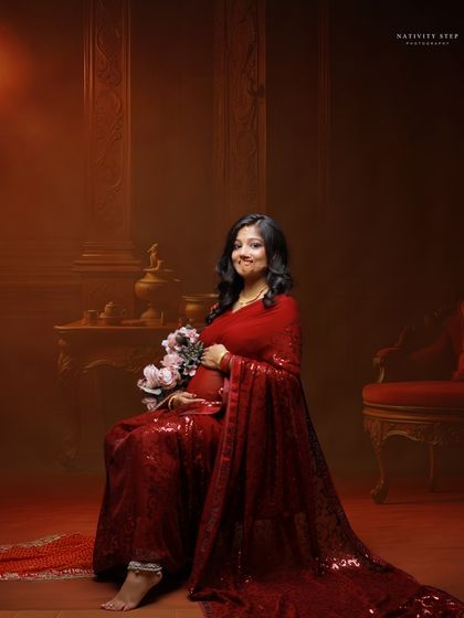 A seated portrait in a shimmering red saree. The warm, dark background and the addition of flowers create a romantic and luxurious feel.