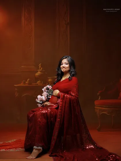 A seated portrait in a shimmering red saree. The warm, dark background and the addition of flowers create a romantic and luxurious feel.