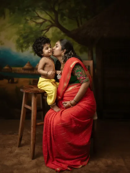 A truly special moment combining a maternity shoot with a cultural theme. Here, an expecting mother shares a tender kiss with her son, who is dressed as Little Krishna. The painted backdrop of a village scene adds a timeless, artistic quality to this story of love.