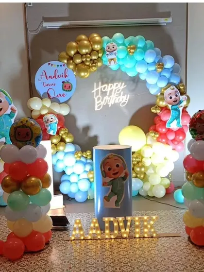 A personalized Cocomelon first birthday party for Aadvik. The decoration includes a circular balloon arch, multiple character cutouts, light-up marquee letters for the name, and a custom sign.