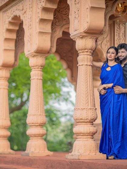 A stylish portrait of a couple in a palace setting. The vibrant blue saree stands out beautifully against the warm tones of the stone pillars.