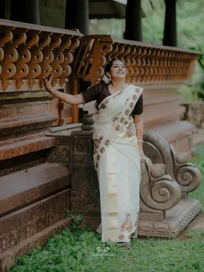 The bride posing against the intricate woodwork of a traditional Kerala building, her joy evident in her smile.