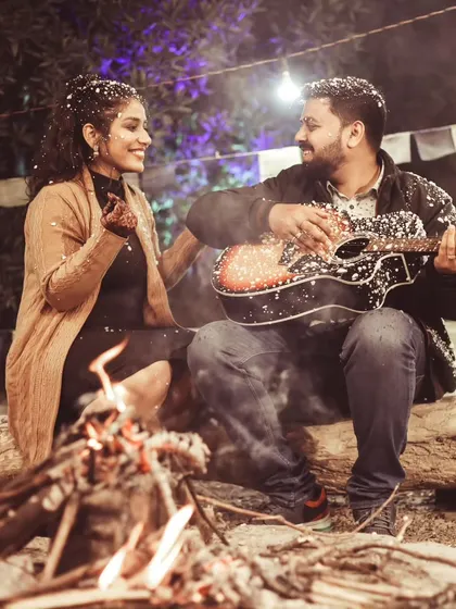 A cozy and romantic campfire scene from a themed pre-wedding shoot. The warm firelight, falling snow effect, and the groom playing a guitar create a magical and intimate atmosphere.