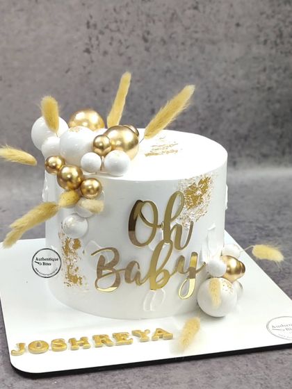 A chic boho-themed "Oh Baby" shower cake. The clean white design is accented with gold and white spheres and dried botanicals for a trendy, rustic look.