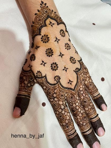 An intricate party henna design with a large, scalloped pattern filled with small floral butis. The fingers are covered in a dense, swirling pattern.
