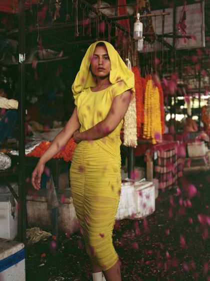 A striking image from the 'Phool Mandi' editorial, featuring a modern, ruched yellow dress with a hood. The falling rose petals add a layer of texture and movement, contrasting the contemporary silhouette with the traditional market setting.