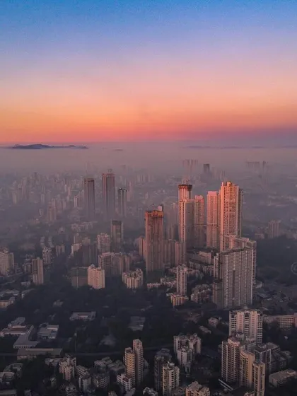 A high-altitude drone shot captures the mountains visible behind Mumbai's hazy, polluted skyline at sunrise.