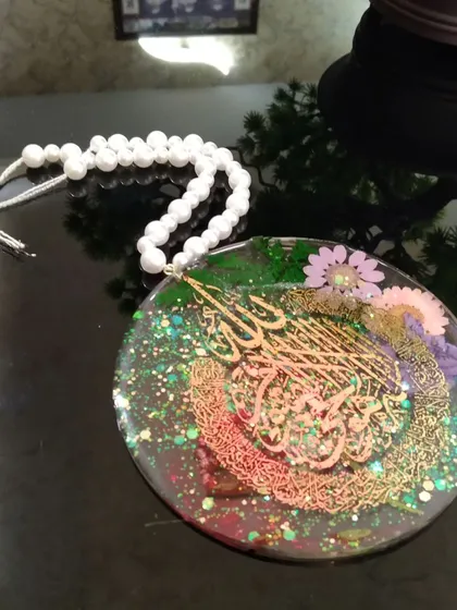 A circular car hanging with Ayatul Kursi calligraphy, embedded with real flowers and glitter, and finished with a pearl tasbih chain.
