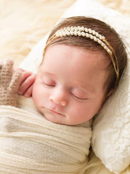 So tiny, so perfect. This close-up shows a newborn sleeping peacefully on a soft pillow, holding a tiny knitted teddy bear. Every detail is captured, from the pearl headband to her tiny fingers.