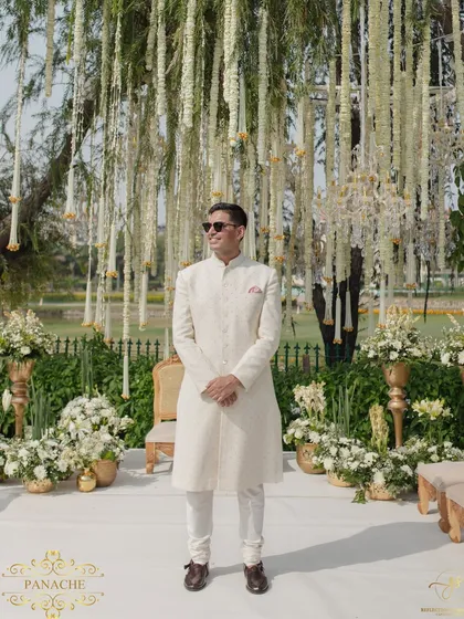 The groom, looking dapper in his white sherwani, waits at the mandap. The backdrop of hanging florals creates a dreamy and dramatic setting.