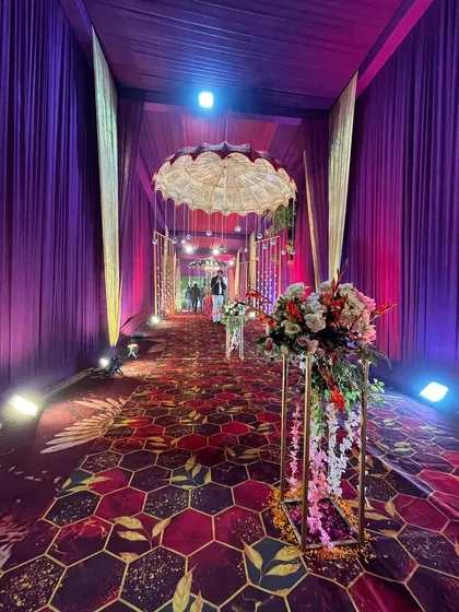 A long, inviting walkway under a canopy of purple drapes, with floral stands and hanging umbrellas guiding guests towards the main celebration area.