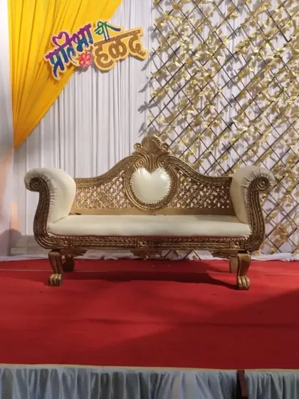 A simple yet elegant Haldi stage. The setup includes a traditional carved sofa against a backdrop of white and yellow drapes and a golden lattice panel, with a custom cutout of the bride's name.