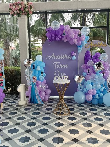 A grand "Frozen" themed first birthday party. This large-scale setup includes custom-printed backdrops, balloon arrangements in shades of purple and blue, and cutouts of Elsa and other characters.