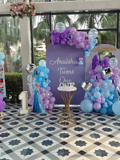 A grand Frozen themed party for Anaisha's first birthday. This elaborate setup includes multiple personalized backdrops, a milestone photo board, character cutouts, and balloon decorations in the signature purple and blue colors.