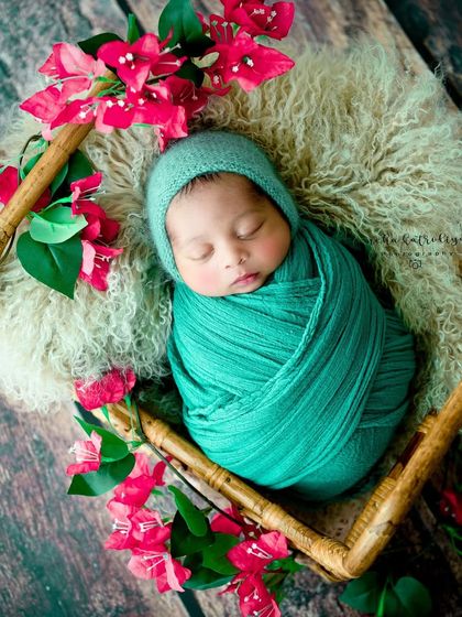 An angled overhead shot showing the baby in a teal wrap, surrounded by bright pink flowers in a unique square basket.