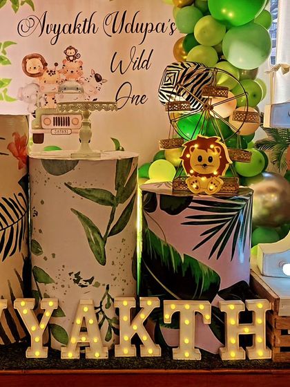 A "Wild One" jungle party setup with a circular backdrop and themed plinths. The decor includes a light-up number '1' and marquee letters spelling out the birthday boy's name.