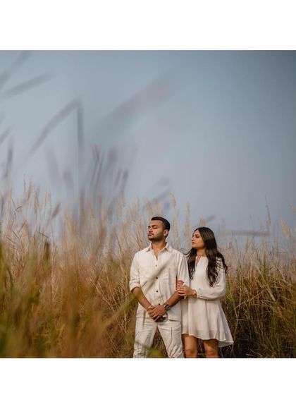 The couple stands together in a field of tall, dry grass. The soft, hazy light and their serene expressions create a dreamy, romantic atmosphere.
