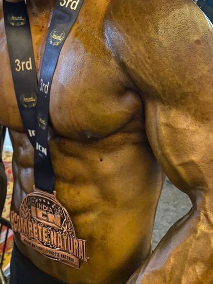 A close-up of a client's physique on show day, wearing his 3rd place medal. The vascularity and grainy muscle detail are signs of peak conditioning.