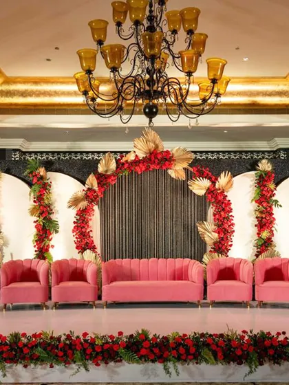 A vibrant and glamorous stage design featuring bold pink seating and a dramatic arch of red roses and gold-painted palm leaves. The ornate chandelier adds a touch of classic luxury to the modern color scheme.