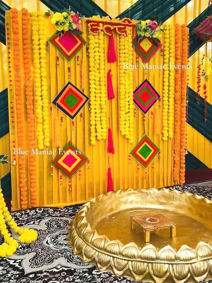 A traditional Haldi setup with a backdrop of yellow and green drapes, adorned with colorful kite-shaped hangings and marigold garlands.