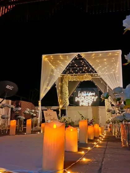 A romantic rooftop proposal scene at night. A pathway lined with large LED candles guides the way to a beautifully lit cabana, framing a "Will you Marry Me?" neon sign for a picture-perfect moment.