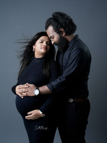 An intimate couple portrait against a solid grey background. The husband's gentle embrace and the wind in his wife's hair create a romantic and emotional moment.