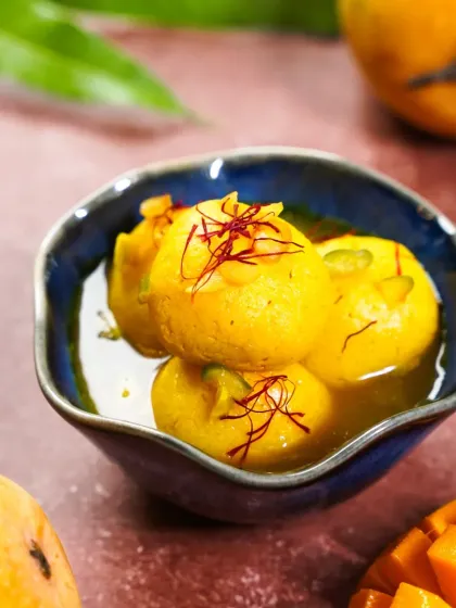 A bowl of luscious Mango Rasgullas soaked in syrup, garnished with saffron. The deep blue ceramic bowl provides a beautiful contrast to the golden yellow of the mangoes.