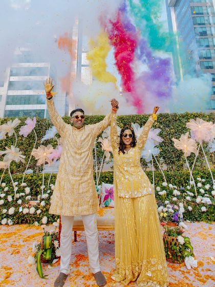 A joyous Haldi moment with the couple celebrating under a rainbow of colored smoke, set against a modern backdrop of whimsical fabric flowers and an urban skyline.