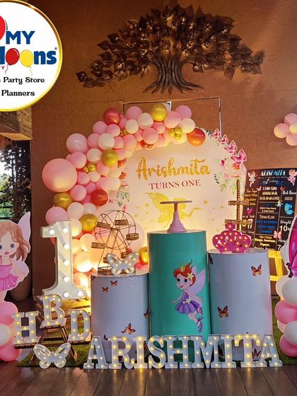 A whimsical fairy-themed first birthday. The pink and gold decor features a 'She leaves a little sparkle' backdrop, fairy cutouts, and a large light-up number '1'.