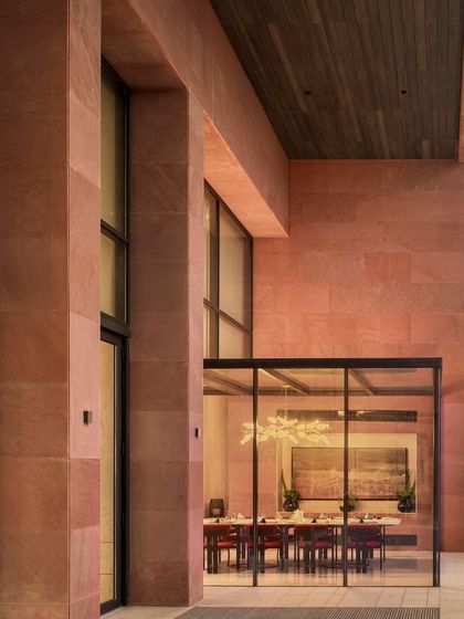 The exterior view of the Elevate Clubhouse at dusk, where the warm interior lighting glows through the large glass walls. The red sandstone facade provides a strong, grounding presence.