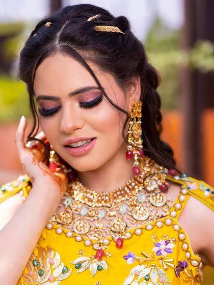 A vibrant and colorful Mehendi look. The makeup features bright pink and purple eyeshadow to match the festive yellow outfit, creating a look that is fun, celebratory, and full of life.