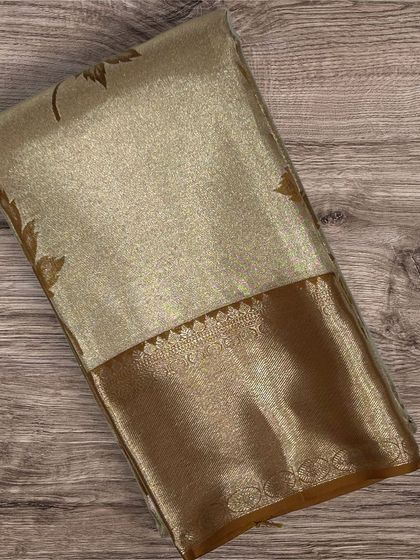 A shimmering gold tissue Kanchi silk saree with a brown border and delicate leaf motifs.