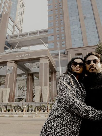 A wide-angle shot capturing a couple in winter coats in front of a modern high-rise building, 'The Collection' at UB City, Bangalore.