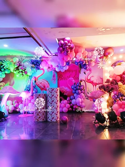 A dynamic and colorful stage setup for the Flamingo party. The decor includes multiple balloon arches, flamingo and mushroom props, and vibrant, multi-colored lighting that creates a fun party atmosphere.