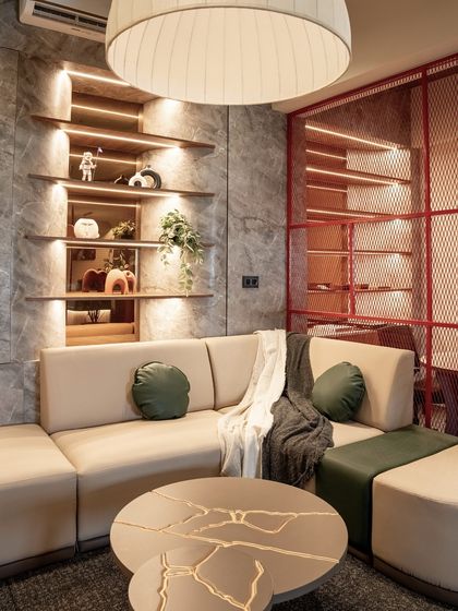 This cozy lounge area within the office provides a space for informal meetings or relaxation. The design features a comfortable sectional sofa, a unique kintsugi-inspired coffee table, and warm, integrated shelving lights.