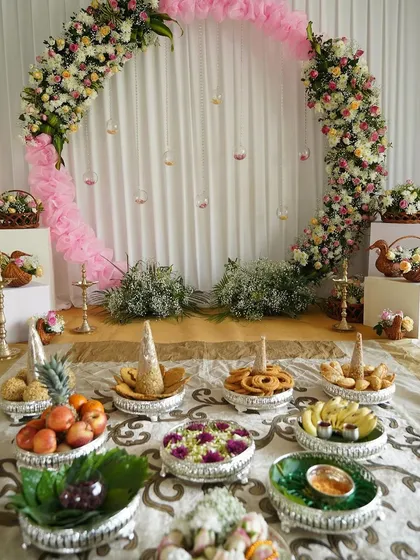 A full view of a half-saree ceremony setup, featuring a floral arch and traditional offerings beautifully arranged on silver platters. The pink fabric accents add a soft, celebratory touch.