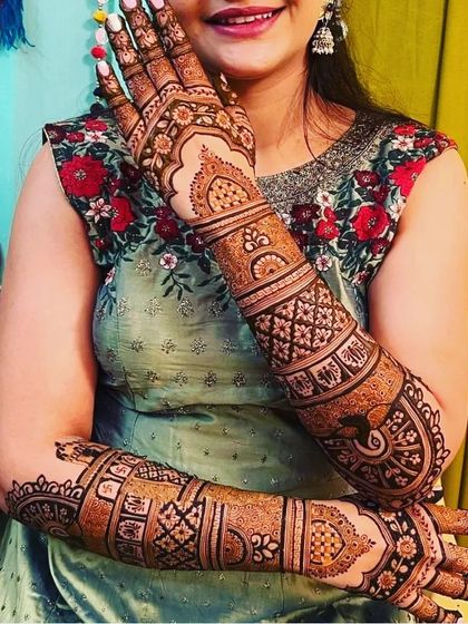 A happy bride showing off her elaborate full-arm mehndi. The design includes classic elements like peacocks and intricate geometric bands.