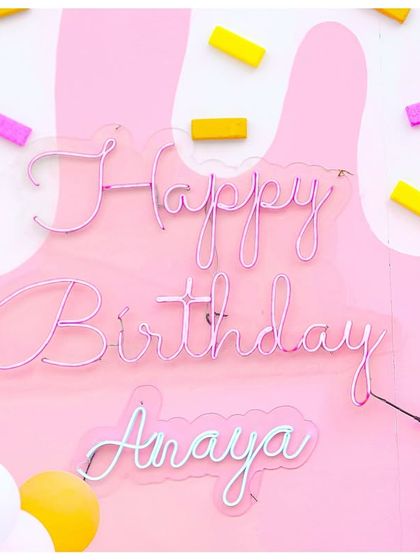 A personalized neon sign for the birthday girl, 'Happy Birthday Anaya', set against a pink backdrop with 3D sprinkles.