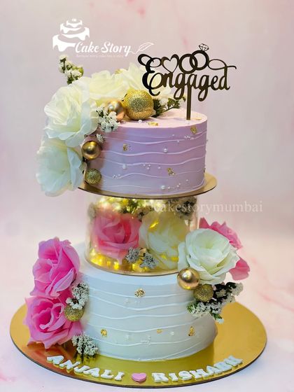 A stunning three-tier engagement cake with a modern design. It features two tiers separated by a clear, light-up spacer, and is decorated with pink and white flowers and gold accents.
